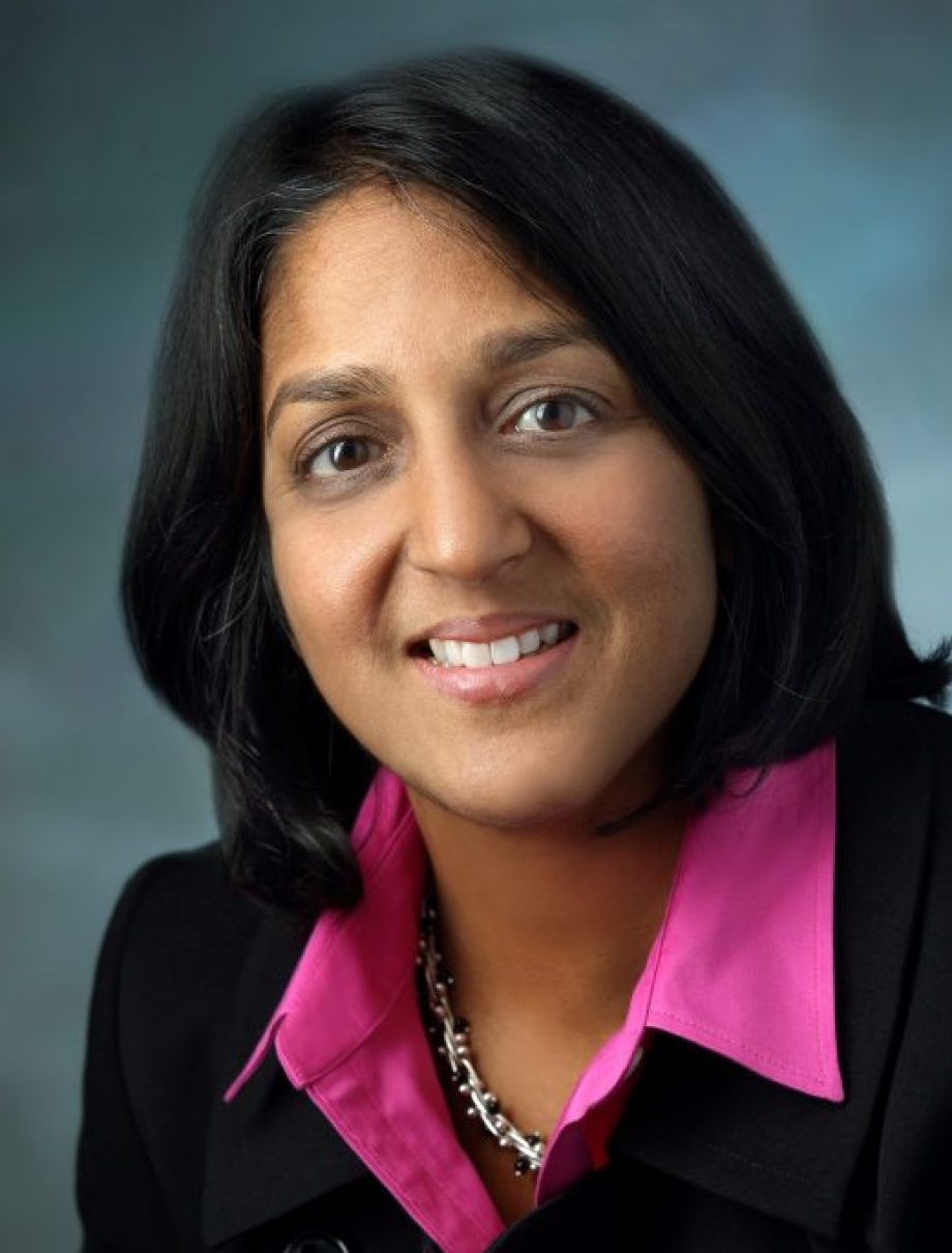Amita Gupta, MD, MHS, is pictured from the shoulders up. She is wearing a bright pink blouse, a black sports jacket, and is posing in front of a blue background. She is smiling.