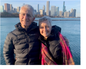 David and Suzanne Thomas pose in an urban waterfront setting. He is wearing a black parka and she is wrapped in a pink plaid shawl. They are smiling.