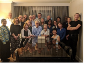 David and Suzanne Thomas are posed at their dining room table, surrounded by a group of Johns Hopkins faculty and infectious diseases fellows. Everyone is smiling.